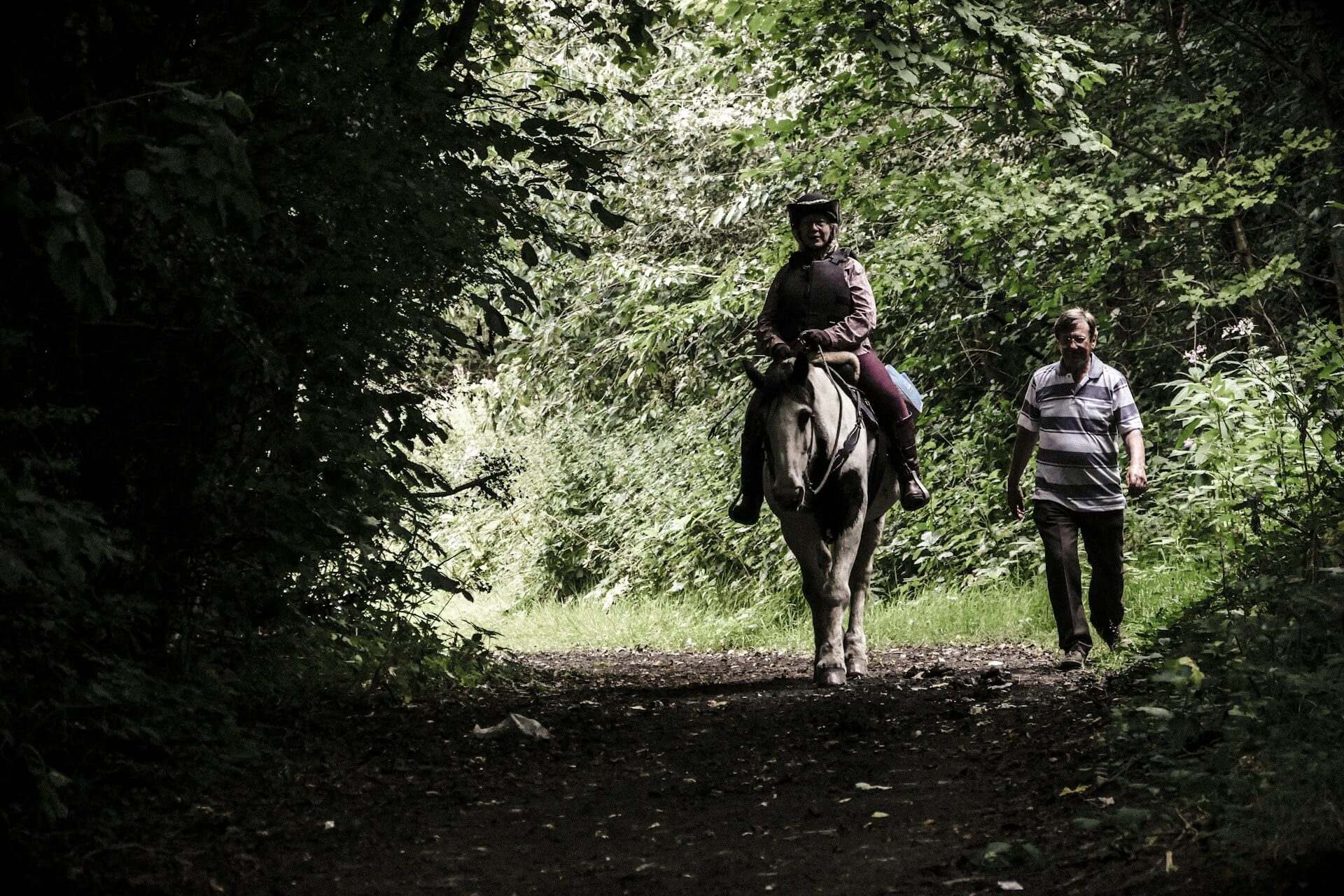 A person riding a horse through the forest, with a man walking beside them