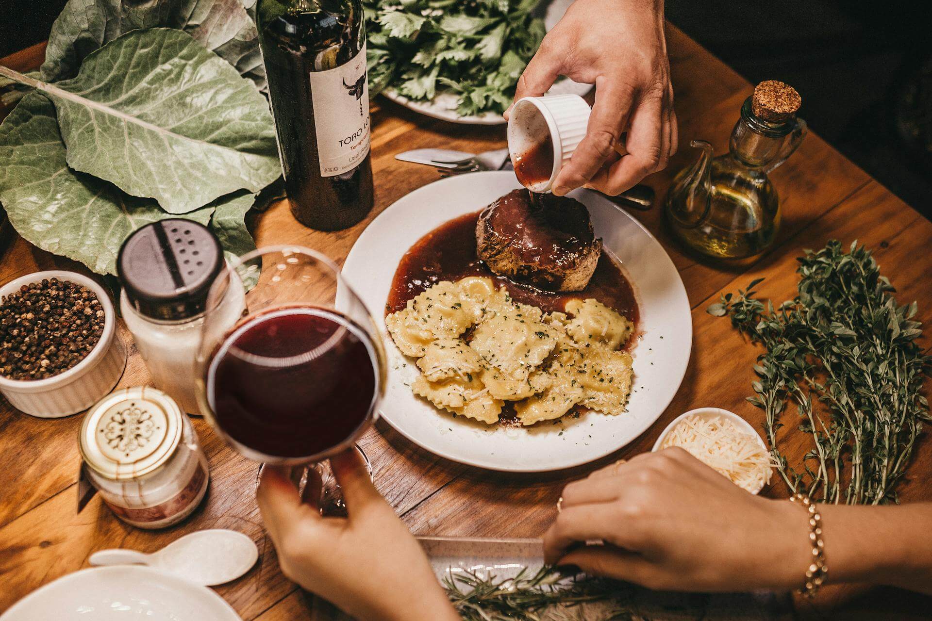 Table with spices and herbs, a person holding a glass of red wine, and another pouring gravy over meat and tortellini